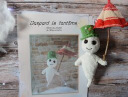 Des pages du patron de Gaspard le fantôme sont posées sur une table en bois. Sur le côté de ces pages, se trouvent une version du fantôme avec son chapeau et son parapluie. De l'autre côté, on aperçoit une guirlande lumineuse, une sorte de nuage blanc et un bout de citrouille.