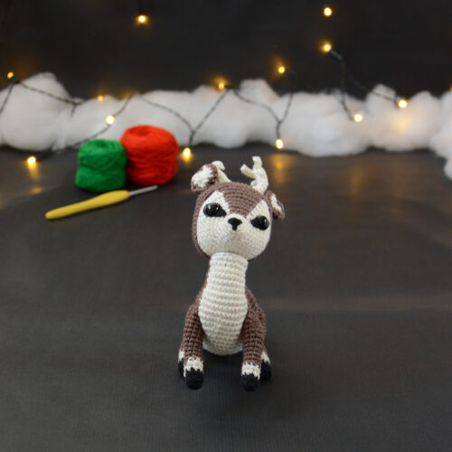 Amigurumi représentant un cerf brun et crème, assis sur l'arrière train, avec des bois crochetés couleur crème. En arrière-plan il y a des pelotes rouge et verte, un crochet ainsi qu'une guirlande lumineuse et des nuages de ouate.