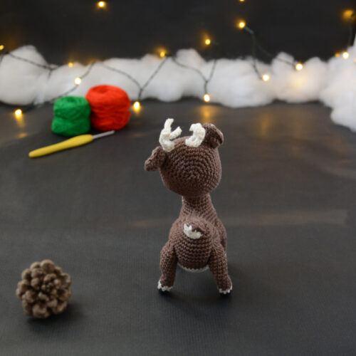 Amigurumi vue de dos représentant un cerf brun et crème avec des bois crochetés couleur crème. Sur l'avant, à gauche se trouve une pomme de pin. En arrière-plan il y a des pelotes rouge et verte, un crochet ainsi qu'une guirlande lumineuse et des nuages de ouate.
