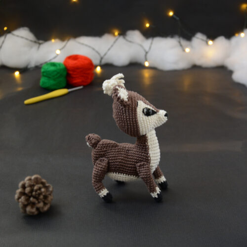 Amigurumi vue de côté représentant un cerf brun et crème avec des bois crochetés couleur crème. Sur l'avant, à gauche se trouve une pomme de pin. En arrière-plan il y a des pelotes rouge et verte, un crochet ainsi qu'une guirlande lumineuse et des nuages de ouate.