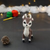 Amigurumi vue de face représentant un cerf brun et crème avec des bois crochetés couleur crème. Sur l'avant, à gauche se trouve une pomme de pin. En arrière-plan il y a des pelotes rouge et verte, un crochet ainsi qu'une guirlande lumineuse et des nuages de ouate.