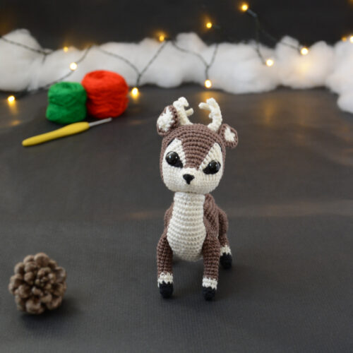 Amigurumi vue de face représentant un cerf brun et crème avec des bois crochetés couleur crème. Sur l'avant, à gauche se trouve une pomme de pin. En arrière-plan il y a des pelotes rouge et verte, un crochet ainsi qu'une guirlande lumineuse et des nuages de ouate.