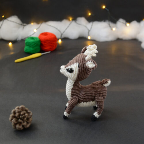 Amigurumi vue de face représentant un cerf brun et crème avec des bois crochetés couleur crème. Sur l'avant, à gauche se trouve une pomme de pin. En arrière-plan il y a des pelotes rouge et verte, un crochet ainsi qu'une guirlande lumineuse et des nuages de ouate.