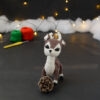 Amigurumi représentant un cerf brun et crème, derrière une pomme de pin, avec des bois crochetés couleur crème. En arrière-plan il y a des pelotes rouge et verte, un crochet ainsi qu'une guirlande lumineuse et des nuages de ouate.