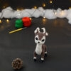 Amigurumi vue de face représentant un cerf brun et crème avec des bois crochetés couleur crème. Sur l'avant, à gauche se trouve une pomme de pin. En arrière-plan il y a des pelotes rouge et verte, un crochet ainsi qu'une guirlande lumineuse et des nuages de ouate.