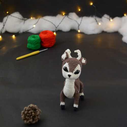 Amigurumi vue de face représentant un cerf brun et crème avec des bois crochetés couleur crème. Sur l'avant, à gauche se trouve une pomme de pin. En arrière-plan il y a des pelotes rouge et verte, un crochet ainsi qu'une guirlande lumineuse et des nuages de ouate.