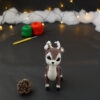 Amigurumi vue de face représentant un cerf brun et crème avec des bois crochetés couleur crème. Sur l'avant, à gauche se trouve une pomme de pin. En arrière-plan il y a des pelotes rouge et verte, un crochet ainsi qu'une guirlande lumineuse et des nuages de ouate.