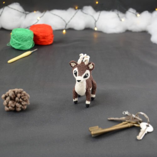 Amigurumi représentant un petit cerf brun et crème avec des bois crochetés couleur crème. Il est un peu tourné sur le côté. Sur la gauche se trouve une pomme de pin. En arrière-plan il y a des pelotes rouge et verte, un crochet ainsi qu'une guirlande lumineuse et des nuages de ouate. Un trousseau de clés est posé au premier plan.