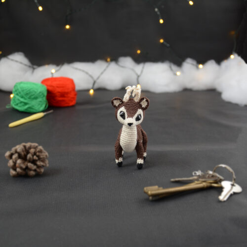Amigurumi représentant un petit cerf brun et crème avec des bois crochetés couleur crème. Sur la gauche se trouve une pomme de pin. En arrière-plan il y a des pelotes rouge et verte, un crochet ainsi qu'une guirlande lumineuse et des nuages de ouate. Un trousseau de clés est posé au premier plan.