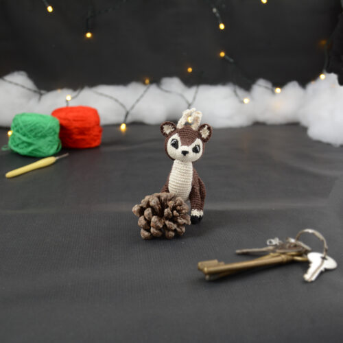 Amigurumi représentant un petit cerf brun et crème, derrière une pomme de pin, avec des bois crochetés couleur crème. En arrière-plan il y a des pelotes rouge et verte, un crochet ainsi qu'une guirlande lumineuse et des nuages de ouate. Un trousseau de clés est posé au premier plan.