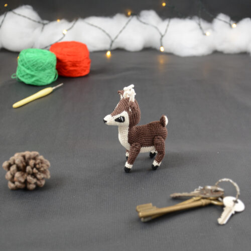 Amigurumi représentant un petit cerf brun et crème, vu de côté, avec des bois crochetés couleur crème. Sur la gauche se trouve une pomme de pin. En arrière-plan il y a des pelotes rouge et verte, un crochet ainsi qu'une guirlande lumineuse et des nuages de ouate. Un trousseau de clés est posé au premier plan.