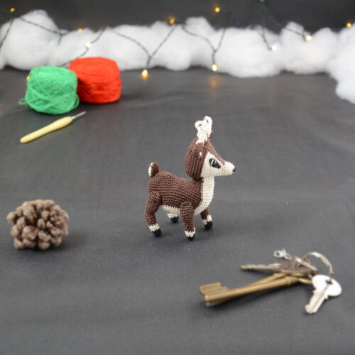 Amigurumi représentant un petit cerf brun et crème, vu de côté, avec des bois crochetés couleur crème. Sur la gauche se trouve une pomme de pin. En arrière-plan il y a des pelotes rouge et verte, un crochet ainsi qu'une guirlande lumineuse et des nuages de ouate. Un trousseau de clés est posé au premier plan.