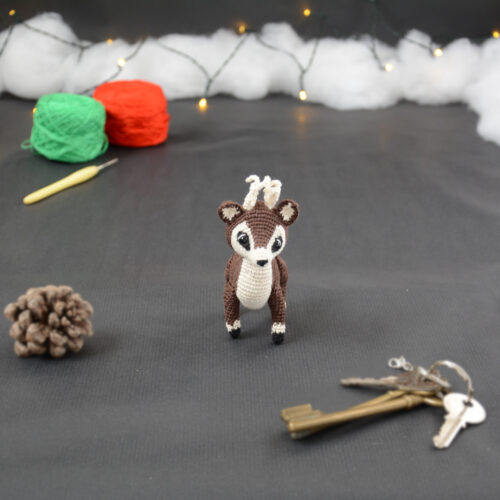 Amigurumi représentant un petit cerf brun et crème avec des bois crochetés couleur crème. Sur la gauche se trouve une pomme de pin. En arrière-plan il y a des pelotes rouge et verte, un crochet ainsi qu'une guirlande lumineuse et des nuages de ouate. Un trousseau de clés est posé au premier plan.