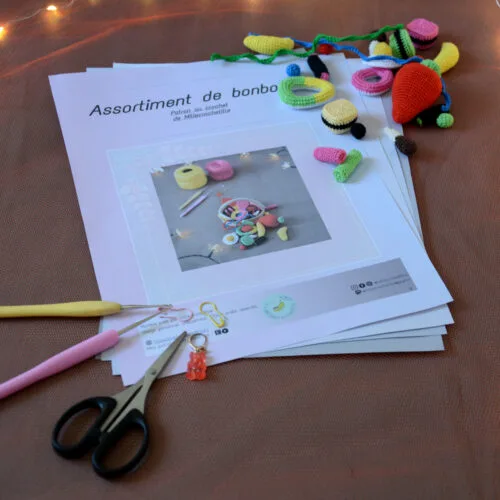Des pages du patron de l'assortiment de bonbons sont posés sur un fond orangé. Dans le coin supérieur droit de ces pages, se trouve différents bonbons (frites, bananes, grosse fraise, réglisses, anneau apollo, bouteille de cola, etc). Dans le coin inférieur gauche sont disposés plusieurs crochets ainsi que des anneaux marqueurs et une paire de ciseaux. À l'arrière-plan on distingue une guirlande lumineuse.