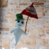 Gaspard le fantôme blanc au crochet portant un chapeau vert. Il tient aussi un parapluie rouge et crème dans sa main. Il se tient devant un mur en pierre.