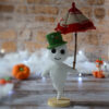 Gaspard le fantôme blanc au crochet, sur un socle en bois. Il porte un chapeau vert et tient un parapluie rouge et crème dans sa main. Il se tient devant un mur en pierre. On distingue en fond flouté, un nuage blanc ainsi qu'une guirlande lumineuse, avec des citrouilles et une oie citrouille posées devant.