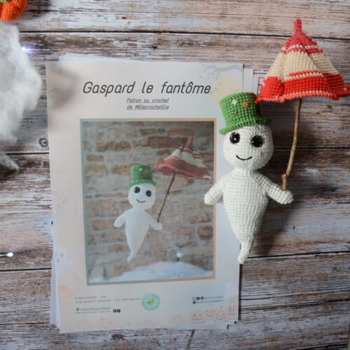 Des pages du patron de Gaspard le fantôme sont posées sur une table en bois. Sur le côté de ces pages, se trouvent une version du fantôme avec son chapeau et son parapluie. De l'autre côté, on aperçoit une guirlande lumineuse, une sorte de nuage blanc et un bout de citrouille.