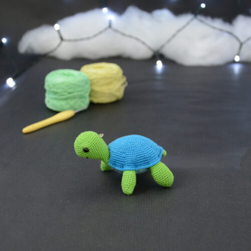 Amigurumi représentant une tortue verte et bleue crochetée, tournée vers la gauche. Elle est posée sur un fond sombre avec en fond, des pelotes de coton vert et jaune ainsi qu’un crochet. En arrière plan, il y a une guirlande lumineuse et un nuage de ouate.