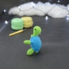 Amigurumi représentant une tortue verte et bleue crochetée, dressée sur ses arrières et tournée vers la droite. Elle est posée sur un fond sombre avec en fond, des pelotes de coton vert et jaune ainsi qu’un crochet. En arrière plan, il y a une guirlande lumineuse et un nuage de ouate.