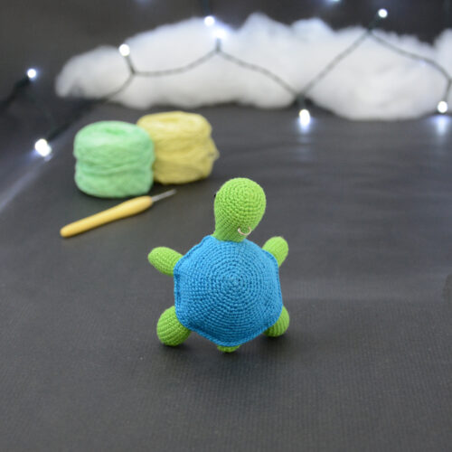 Amigurumi représentant une tortue verte et bleue crochetée, dressée de dos sur ses arrières. Elle est posée sur un fond sombre avec en fond, des pelotes de coton vert et jaune ainsi qu’un crochet. En arrière plan, il y a une guirlande lumineuse et un nuage de ouate.
