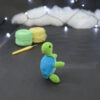 Amigurumi représentant une tortue verte et bleue crochetée, dressée sur ses arrières et tournée vers la droite. Elle est posée sur un fond sombre avec en fond, des pelotes de coton vert et jaune ainsi qu’un crochet. En arrière plan, il y a une guirlande lumineuse et un nuage de ouate.