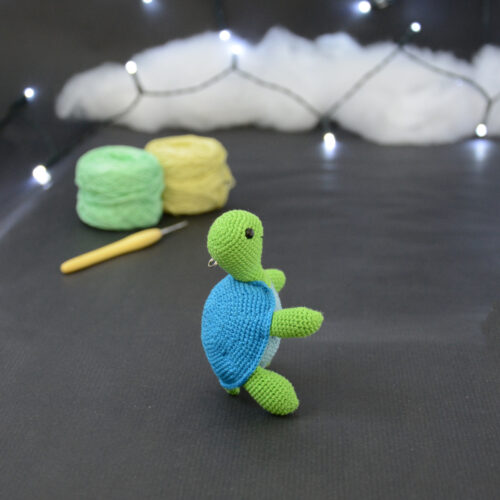 Amigurumi représentant une tortue verte et bleue crochetée, dressée sur ses arrières et tournée vers la droite. Elle est posée sur un fond sombre avec en fond, des pelotes de coton vert et jaune ainsi qu’un crochet. En arrière plan, il y a une guirlande lumineuse et un nuage de ouate.