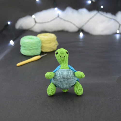 Amigurumi représentant une tortue verte et bleue crochetée, dressée sur ses arrières. Elle est posée sur un fond sombre avec en fond, des pelotes de coton vert et jaune ainsi qu’un crochet. En arrière plan, il y a une guirlande lumineuse et un nuage de ouate.