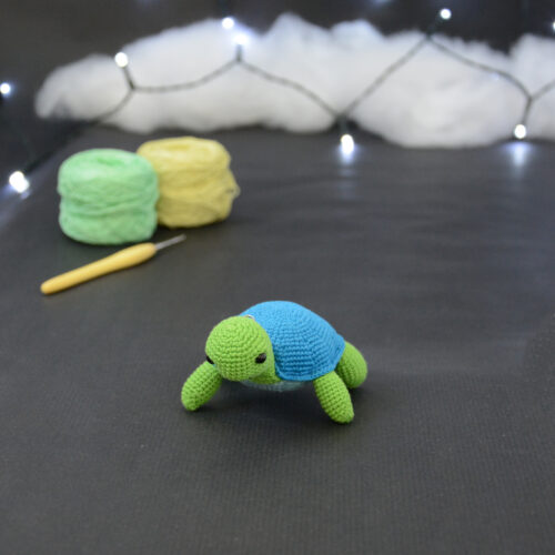 Amigurumi représentant une tortue verte et bleue crochetée, tournée légèrement vers la gauche. Elle est posée sur un fond sombre avec en fond, des pelotes de coton vert et jaune ainsi qu’un crochet. En arrière plan, il y a une guirlande lumineuse et un nuage de ouate.
