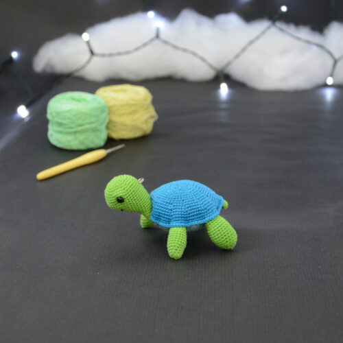 Amigurumi représentant une tortue verte et bleue crochetée, tournée vers la gauche. Elle est posée sur un fond sombre avec en fond, des pelotes de coton vert et jaune ainsi qu’un crochet. En arrière plan, il y a une guirlande lumineuse et un nuage de ouate.