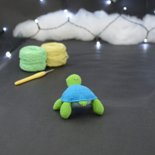 Amigurumi représentant une tortue verte et bleue crochetée, de dos. Elle est posée sur un fond sombre avec en fond, des pelotes de coton vert et jaune ainsi qu’un crochet. En arrière plan, il y a une guirlande lumineuse et un nuage de ouate.