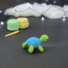 Amigurumi représentant une tortue verte et bleue crochetée, tournée vers la droite. Elle est posée sur un fond sombre avec en fond, des pelotes de coton vert et jaune ainsi qu’un crochet. En arrière plan, il y a une guirlande lumineuse et un nuage de ouate.