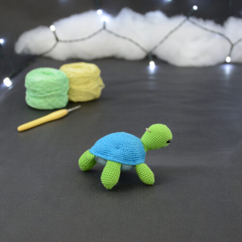 Amigurumi représentant une tortue verte et bleue crochetée, tournée vers la droite. Elle est posée sur un fond sombre avec en fond, des pelotes de coton vert et jaune ainsi qu’un crochet. En arrière plan, il y a une guirlande lumineuse et un nuage de ouate.