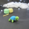 Amigurumi représentant une tortue verte et bleue crochetée, légèrement tournée vers la droite. Elle est posée sur un fond sombre avec en fond, des pelotes de coton vert et jaune ainsi qu’un crochet. En arrière plan, il y a une guirlande lumineuse et un nuage de ouate.