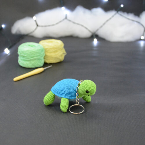 Amigurumi représentant une tortue verte et bleue crochetée, légèrement tournée vers la droite. Un porte-clefs retombe sur le côté de son cou. Elle est posée sur un fond sombre avec en fond, des pelotes de coton vert et jaune ainsi qu’un crochet. En arrière plan, il y a une guirlande lumineuse et un nuage de ouate.