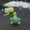 Amigurumi représentant une tortue verte et bleue crochetée, dressée sur ses arrières. Un porte-clefs retombe sur le devant de son plastron. Elle est posée sur un fond sombre avec en fond, des pelotes de coton vert et jaune ainsi qu’un crochet. En arrière plan, il y a une guirlande lumineuse et un nuage de ouate.