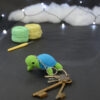 Amigurumi représentant une tortue verte et bleue crochetée, tournée légèrement vers la gauche. Des clefs accrochées à un porte-clefs retombe sur le côté droit de la tortue. Elle est posée sur un fond sombre avec en fond, des pelotes de coton vert et jaune ainsi qu’un crochet. En arrière plan, il y a une guirlande lumineuse et un nuage de ouate.