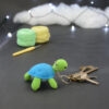 Amigurumi représentant une tortue verte et bleue crochetée, de dos, tournée légèrement vers la droite. Des clefs accrochées à un porte-clefs retombe sur le côté droit de la tortue. Elle est posée sur un fond sombre avec en fond, des pelotes de coton vert et jaune ainsi qu’un crochet. En arrière plan, il y a une guirlande lumineuse et un nuage de ouate.