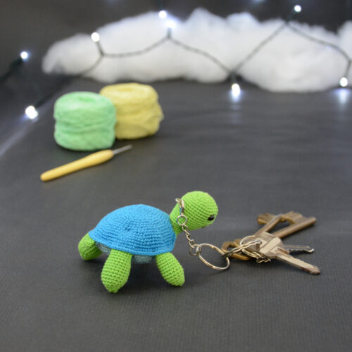 Amigurumi représentant une tortue verte et bleue crochetée, de dos, tournée légèrement vers la droite. Des clefs accrochées à un porte-clefs retombe sur le côté droit de la tortue. Elle est posée sur un fond sombre avec en fond, des pelotes de coton vert et jaune ainsi qu’un crochet. En arrière plan, il y a une guirlande lumineuse et un nuage de ouate.