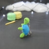 Amigurumi représentant une tortue verte et bleue crochetée, dressée sur ses arrières et tournée vers la gauche. Elle est posée sur un fond sombre avec en fond, des pelotes de coton vert et jaune ainsi qu’un crochet. En arrière plan, il y a une guirlande lumineuse et un nuage de ouate.