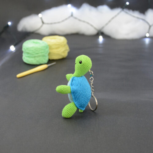 Amigurumi représentant une tortue verte et bleue crochetée, dressée sur ses arrières et tournée vers la gauche. Elle est posée sur un fond sombre avec en fond, des pelotes de coton vert et jaune ainsi qu’un crochet. En arrière plan, il y a une guirlande lumineuse et un nuage de ouate.
