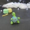 Amigurumi représentant une tortue verte et bleue crochetée, dressée sur ses arrières et légèrement tournée vers la droite. Elle est posée sur un fond sombre avec en fond, des pelotes de coton vert et jaune ainsi qu’un crochet. En arrière plan, il y a une guirlande lumineuse et un nuage de ouate.