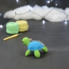 Amigurumi représentant une tortue verte et bleue crochetée, de dos et légèrement tournée vers la gauche. On distingue un porte-clefs sur son dos. Elle est posée sur un fond sombre avec en fond, des pelotes de coton vert et jaune ainsi qu’un crochet. En arrière plan, il y a une guirlande lumineuse et un nuage de ouate.