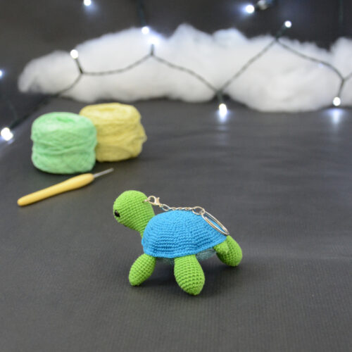 Amigurumi représentant une tortue verte et bleue crochetée, de dos et légèrement tournée vers la gauche. On distingue un porte-clefs sur son dos. Elle est posée sur un fond sombre avec en fond, des pelotes de coton vert et jaune ainsi qu’un crochet. En arrière plan, il y a une guirlande lumineuse et un nuage de ouate.