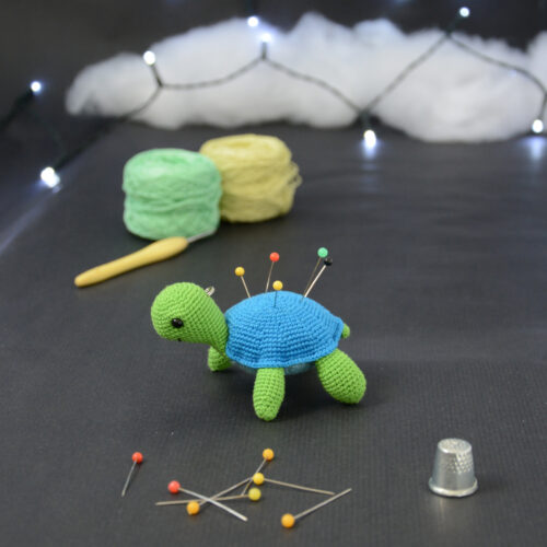 Amigurumi représentant une tortue verte et bleue crochetée, tournée vers la gauche. Elle a des épingles piquées dans sa carapace. Elle est posée sur un fond sombre avec en fond, des pelotes de coton vert et jaune ainsi qu’un crochet. En arrière plan, il y a une guirlande lumineuse et un nuage de ouate. À l'avant-plan sont disposés d'autres épingles ainsi qu'un dés à coudre.