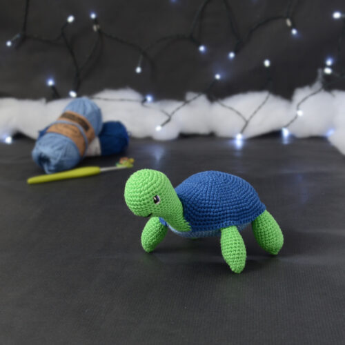 Amigurumi représentant une tortue verte à carapace bleue, tournée vers la gauche, posée sur un fond noir avec des pelotes bleues, un crochet et une guirlande lumineuse en arrière-plan.