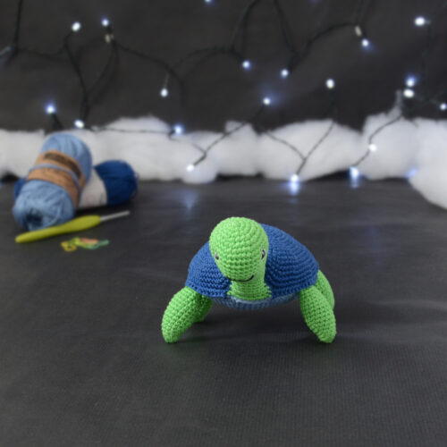 Amigurumi représentant une tortue verte à carapace bleue, vue de face, posée sur un fond noir avec des pelotes bleues, un crochet et une guirlande lumineuse en arrière-plan.