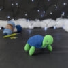 Amigurumi représentant une tortue verte à carapace bleue, tournée vers la droite, posée sur un fond noir avec des pelotes bleues, un crochet et une guirlande lumineuse en arrière-plan.
