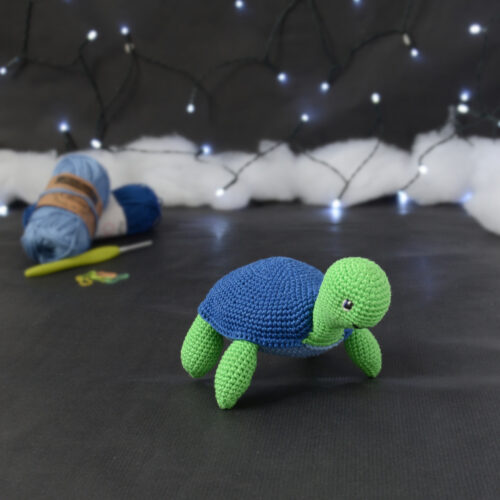 Amigurumi représentant une tortue verte à carapace bleue, tournée vers la droite, posée sur un fond noir avec des pelotes bleues, un crochet et une guirlande lumineuse en arrière-plan.