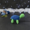 Amigurumi représentant une tortue verte à carapace bleue, de profil orientée vers la droite, posée sur un fond noir avec des pelotes bleues, un crochet et une guirlande lumineuse en arrière-plan.