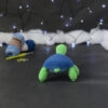 Amigurumi représentant une tortue verte à carapace bleue, vue de dos, posée sur un fond noir avec des pelotes bleues, un crochet et une guirlande lumineuse en arrière-plan.